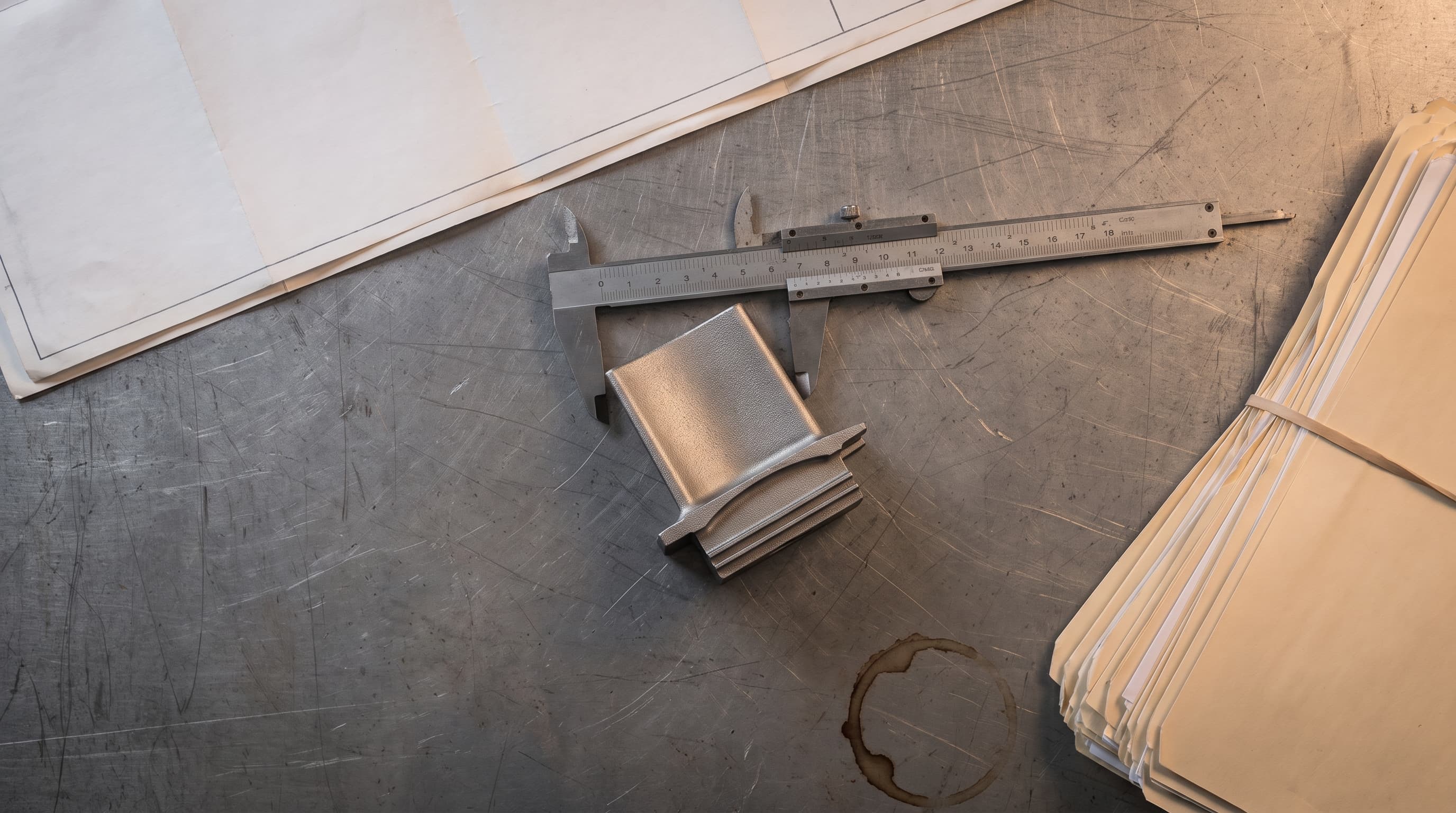 Overhead view of a titanium turbine blade casting being measured with steel calipers beside a stack of unmarked contract folders on a workbench.