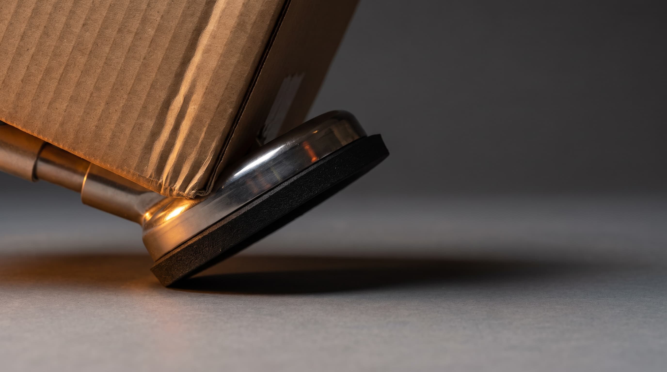 A foam-sealed vacuum gripper pressing against the corner of a cardboard shipping case on a grey seamless surface, lit by a single warm light with a long shadow.